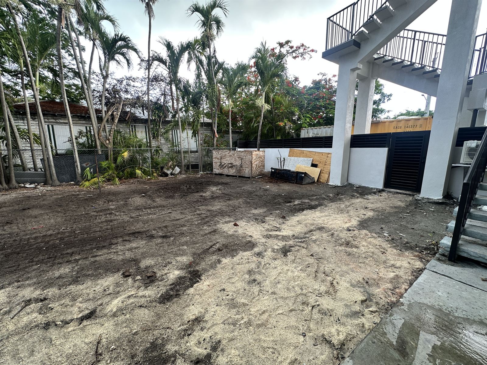 Bare backyard before landscaping — graded sand and soil beneath tall palms, with construction materials along the fence